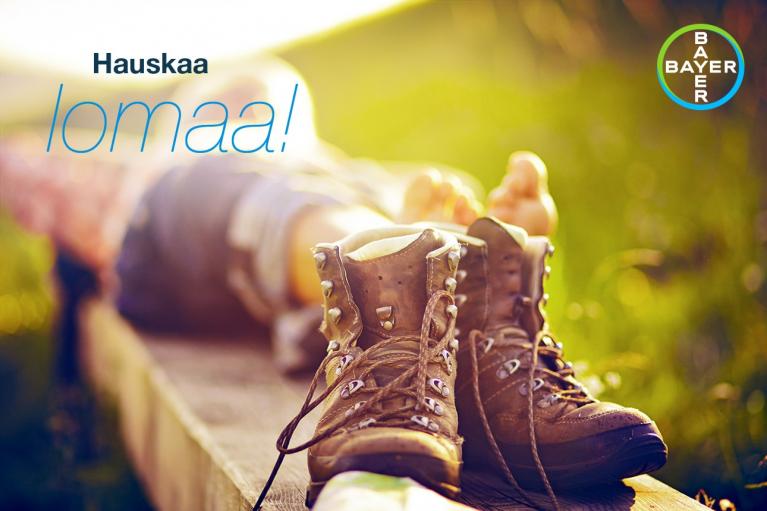 A pair of hiking boots on a wooden surface with a blurred background of feet relaxing. Text reads "Hauskaa lomaa!" with a Bayer logo in the corner.