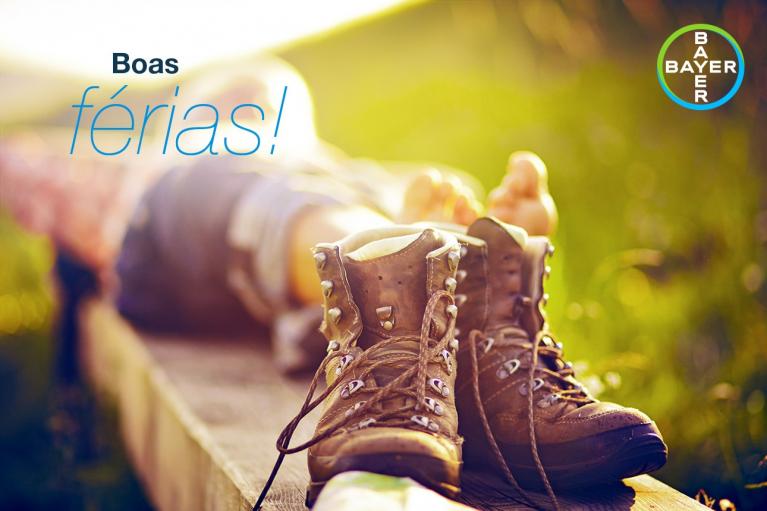 A pair of hiking boots on a wooden plank, with a person relaxing in the background. Text reads "Boas férias!" and features the Bayer logo.