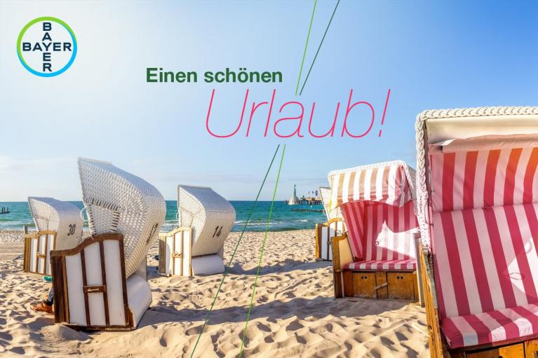 Sandy beach with wicker beach chairs under a clear blue sky, featuring the text "Einen schönen Urlaub!" and a Bayer logo.