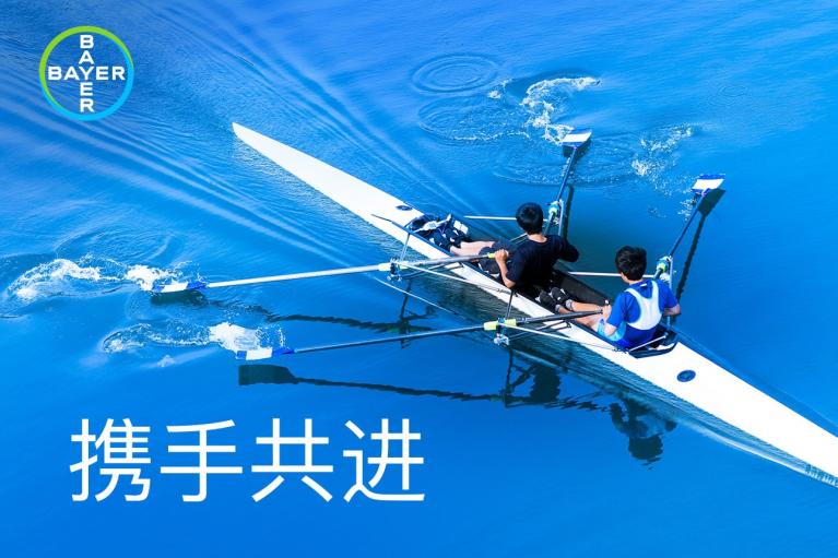 Two people rowing in a sleek white boat on blue water. Bayer logo in the corner. Chinese characters translate to "Join Hands and Move Forward.