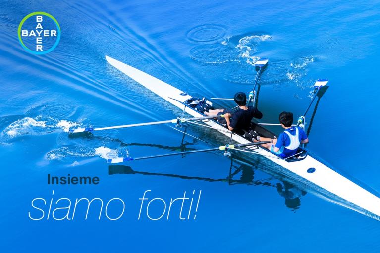 Two people rowing in a blue boat on calm water with the Bayer logo. Italian text reads "Insieme siamo forti!" (Together we are strong!).