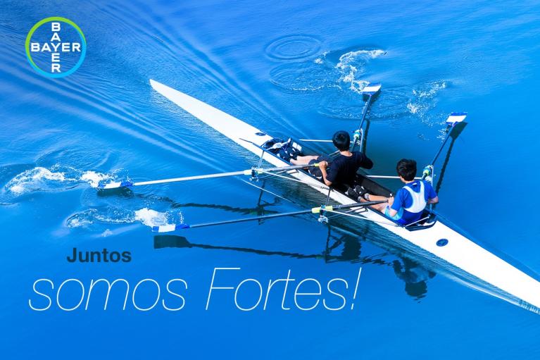 Two rowers in a sleek, white boat glide on calm blue water. Text reads "Juntos somos Fortes!" The Bayer logo is in the top left corner.