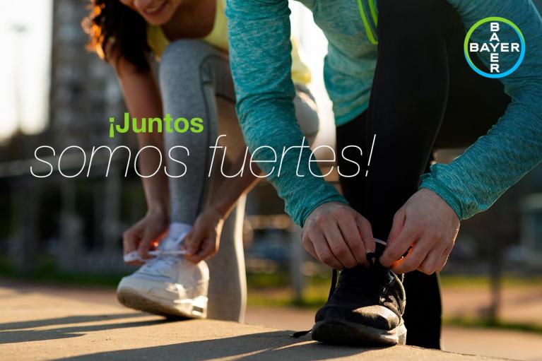 Two people tying shoelaces. Text overlay reads, "¡Juntos somos fuertes!" and the Bayer logo is visible in the corner.