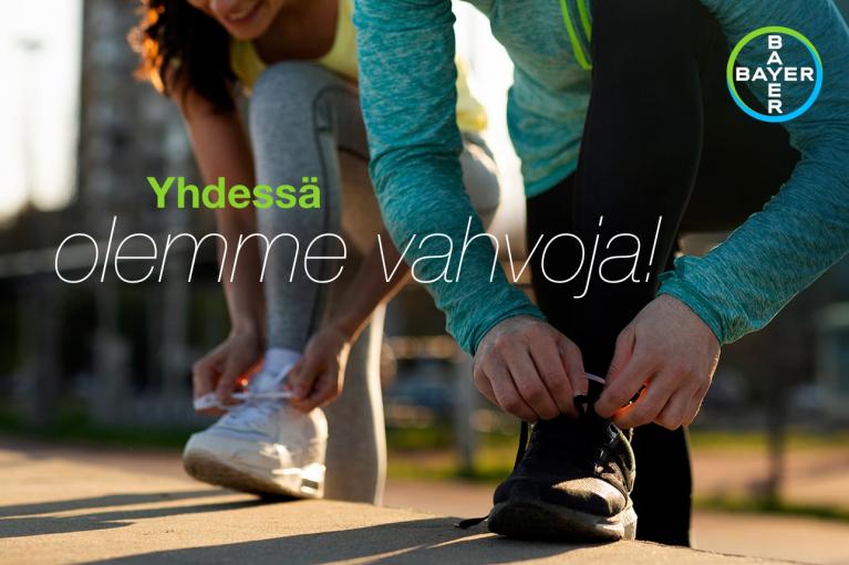 Two people tying their athletic shoes on a sunny outdoor platform. Text reads, "Yhdessä olemme vahvoja!" with a Bayer logo in the corner.