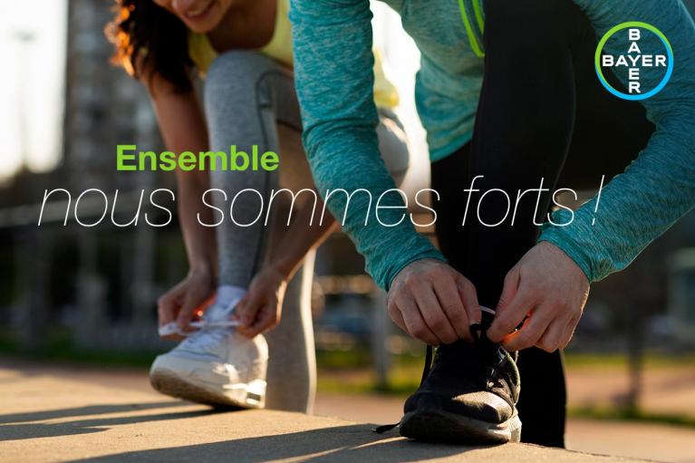 Two people tying their shoelaces, one wearing white sneakers and the other black. Text overlay in French reads, "Ensemble nous sommes forts!" and a Bayer logo is visible.