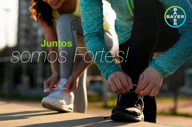 Two people tying their sneakers outdoors with the text "Juntos somos fortes!" and a Bayer logo in the corner.