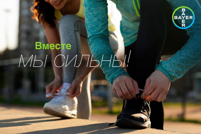 Two people are tying their shoelaces outdoors. Text says, "Вместе мы сильны!" and a Bayer logo is visible.