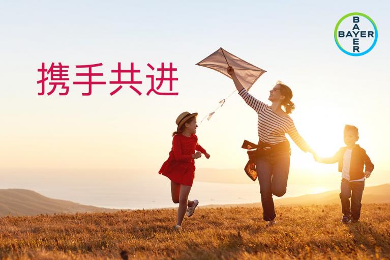 A woman and two children run in a field with a kite at sunset. The Bayer logo is in the top right corner. Chinese text on the left reads "携手共进" (Progress Together).