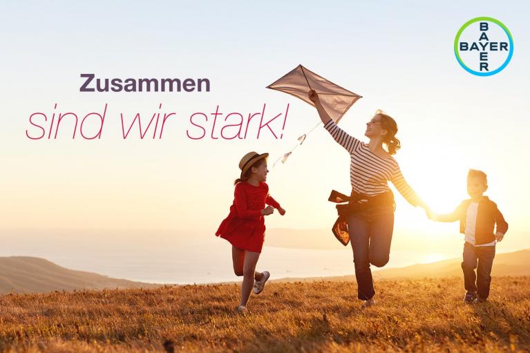 A woman runs with two children on a grassy hill at sunset, holding a kite. The text reads "Zusammen sind wir stark!" with a Bayer logo in the corner.
