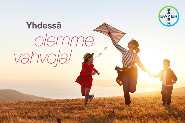 A woman and two children run with a kite in a field at sunset, with text in Finnish and the Bayer logo visible.