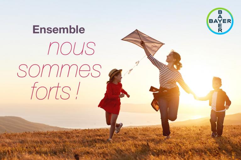 A woman and two children fly a kite on a grassy hill at sunset. Text reads "Ensemble nous sommes forts!" and displays the Bayer logo.