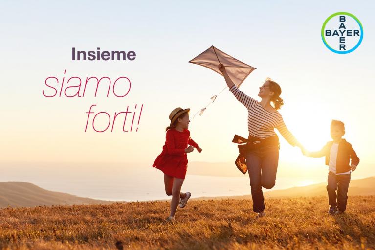 A woman and two children fly a kite on a grassy hill at sunset. The text "Insieme siamo forti!" and Bayer logo are visible.