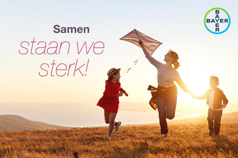 A woman flies a kite with two children on a grassy hill at sunset, with the text "Samen staan we sterk!" and the Bayer logo in the corner.