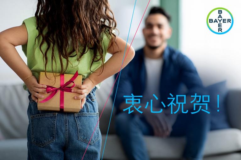 Child holding a wrapped gift behind their back, facing an adult on a couch. Text in Mandarin: "衷心祝贺!" and Bayer logo in the corner.