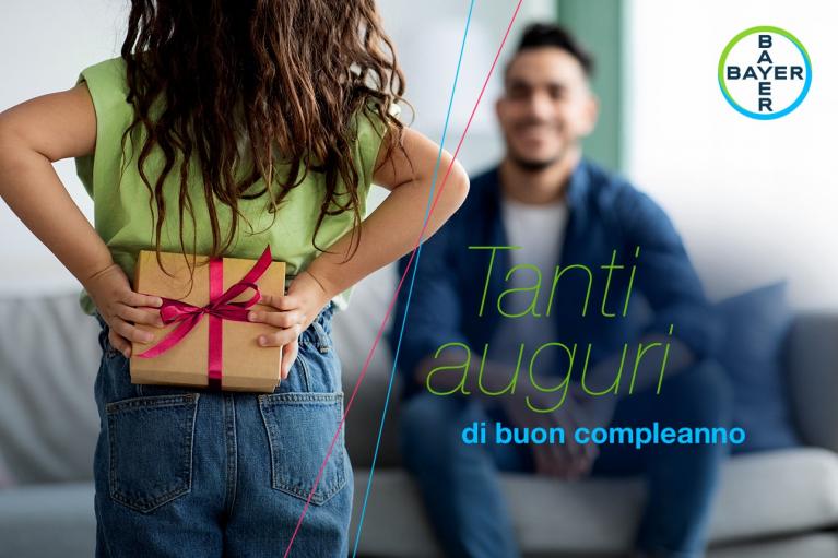 Child hiding a gift behind their back, facing an adult sitting on a couch. Text reads "Tanti auguri di buon compleanno" with Bayer logo in the corner.