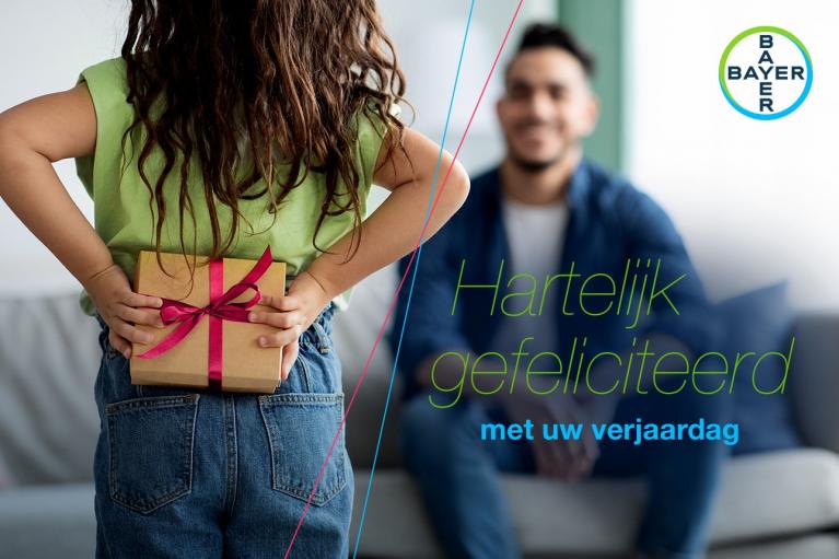 Child holding a gift behind their back, facing a smiling adult on a couch. Dutch text reads "Hartelijk gefeliciteerd met uw verjaardag" with a Bayer logo.