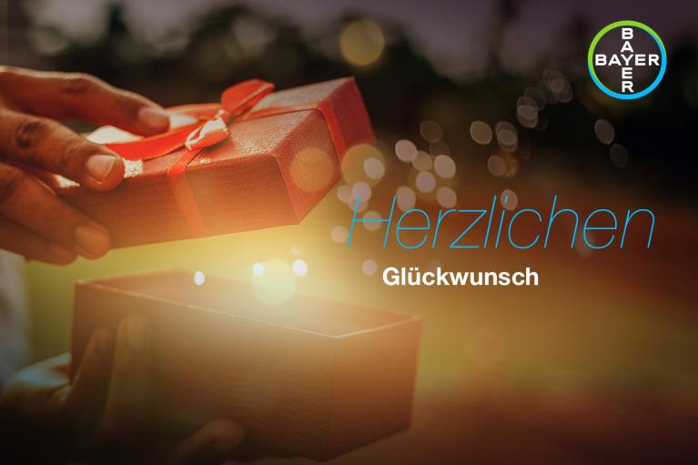 Hands opening a red gift box with a ribbon, surrounded by glowing lights. "Herzlichen Glückwunsch" text and a Bayer logo are visible.