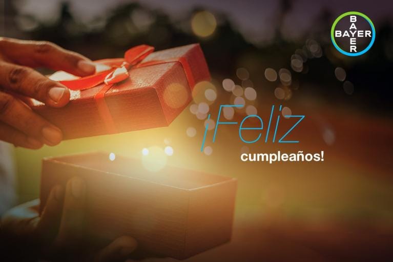 Hands hold an open gift box with "¡Feliz cumpleaños!" text and Bayer logo in the corner.