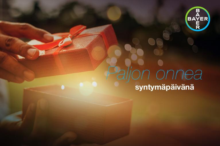Hands opening a gift box with a red ribbon, with the text "Paljon onnea syntymäpäivänä" and a Bayer logo in the corner.