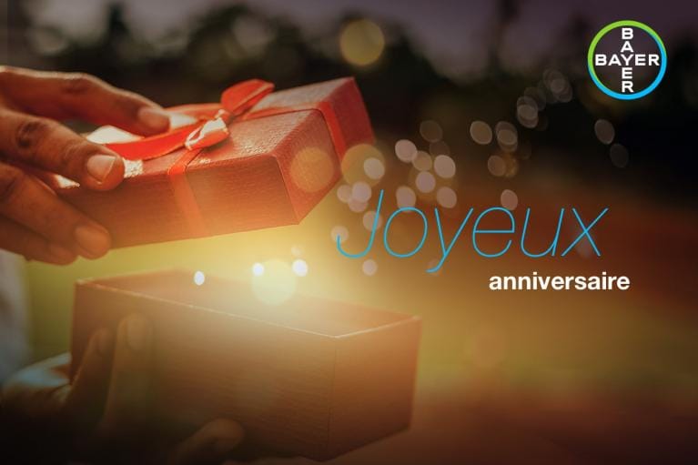 Hands holding an open gift box with glowing light inside. Text reads "Joyeux anniversaire" with a Bayer logo in the top right corner.