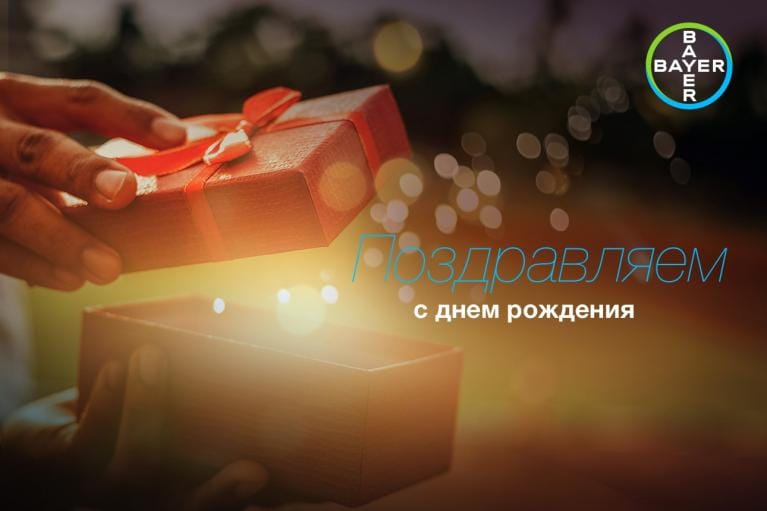 A person opens a red gift box with a ribbon. Russian text reads "Поздравляем с днем рождения," meaning "Happy Birthday." The Bayer logo is in the top right corner.