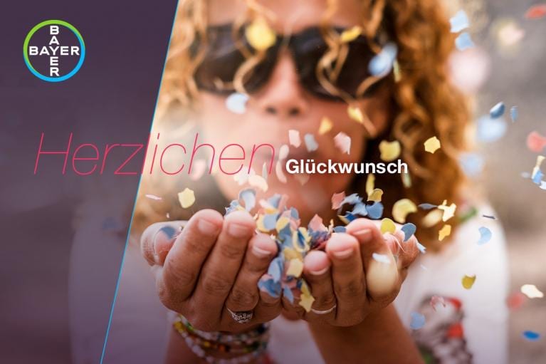 A person blowing colorful confetti from their hands, with the word "Herzlichen Glückwunsch" and the Bayer logo in the corner.