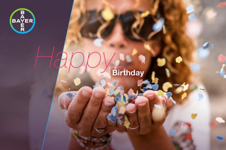 A person with curly hair and sunglasses blows colorful confetti from their hands. The words "Happy Birthday" and a Bayer logo are on the image.