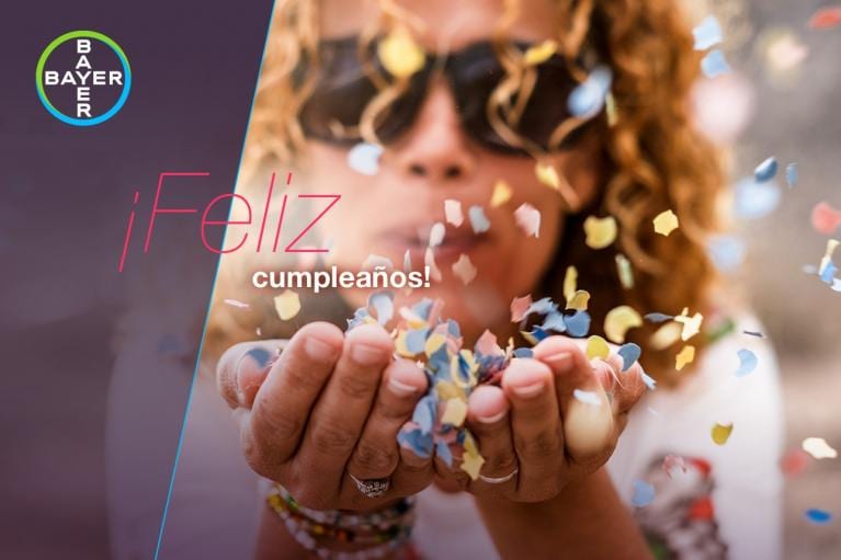 Person blowing colorful confetti towards the camera with "¡Feliz cumpleaños!" text overlay and Bayer logo in the corner.