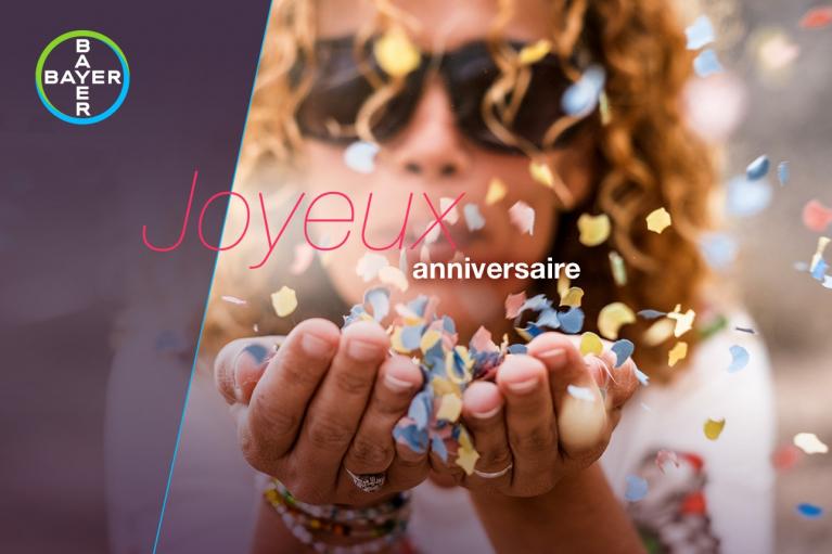 A person with curly hair blowing colorful confetti from cupped hands. The French text reads "Joyeux anniversaire" with a Bayer logo in the corner.