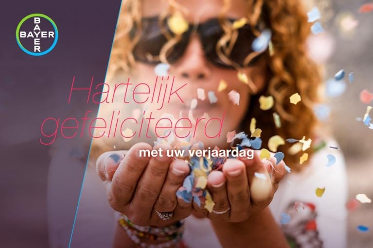 Person blowing colorful confetti from their hands, with "Hartelijk gefeliciteerd met uw verjaardag" text overlay. Bayer logo in the top left corner.