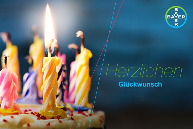 Colorful candles on a cake are lit, with "Herzlichen Glückwunsch" written in German. The Bayer logo appears in the corner.