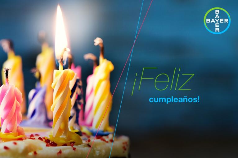 Colorful candles on a cake are lit, with a "Feliz cumpleaños" message and a Bayer logo in the background.
