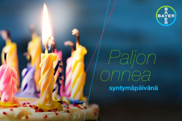 A cake with colorful spiral candles lit on top. The text on the image reads "Paljon onnea syntymäpäivänä" next to a Bayer logo.