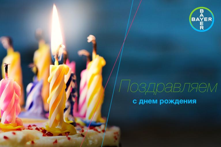 Lit birthday candles on a cake with the Bayer logo and Russian text saying "Congratulations on your birthday.
