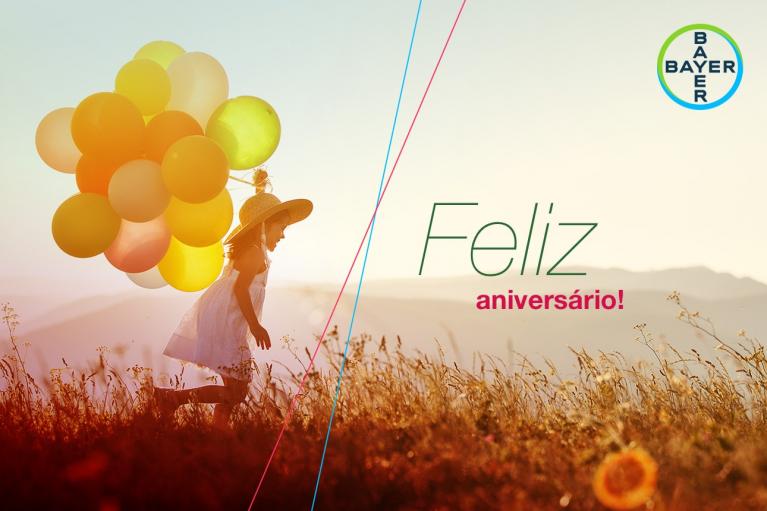 Child with balloons running in a meadow at sunset, with "Feliz aniversário!" text and Bayer logo.