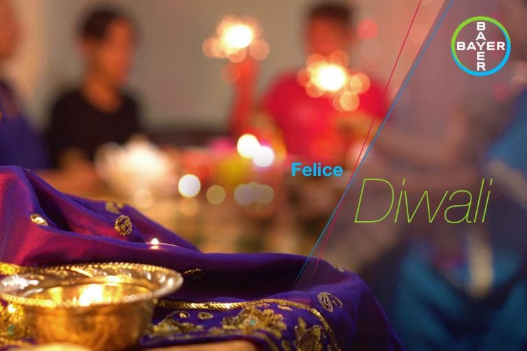 A festive Diwali setting with candles and decorative cloth, framed by the Bayer logo and text reading "Felice Diwali.