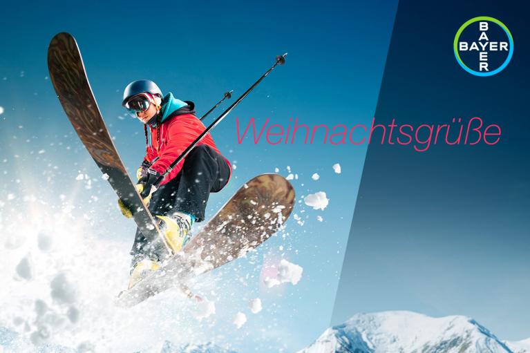 A skier in mid-air against a snowy mountain backdrop with "Weihnachtsgrüße" and a logo in the corner.