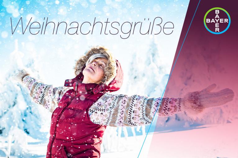 Person in winter clothing stands in snow with arms outstretched, smiling. Text says "Weihnachtsgrüße" with a Bayer logo in the corner.