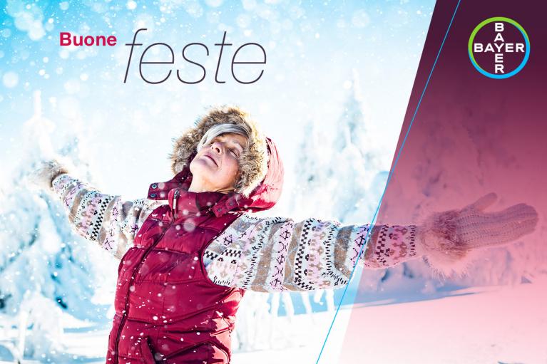Person in a winter scene wearing a red vest and knit sweater, arms outstretched, with "Buone feste" text and Bayer logo in the corner.