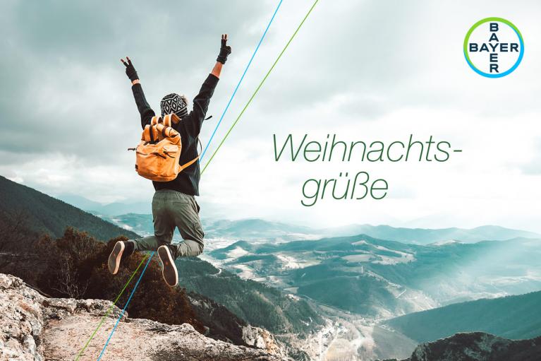 Person with a yellow backpack jumps joyfully on a mountain summit, overlooking a valley. The sky is overcast. Text reads "Weihnachtsgrüße" alongside a Bayer logo.
