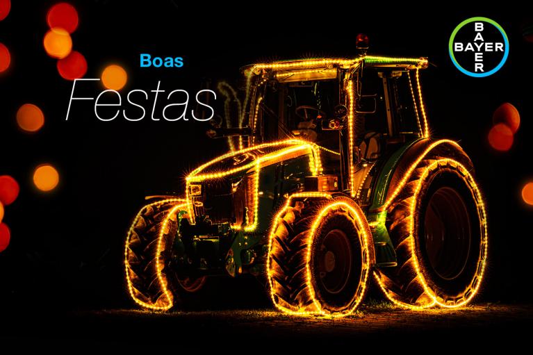A tractor is decorated with bright lights against a dark background. The words "Boas Festas" are visible alongside the Bayer logo.