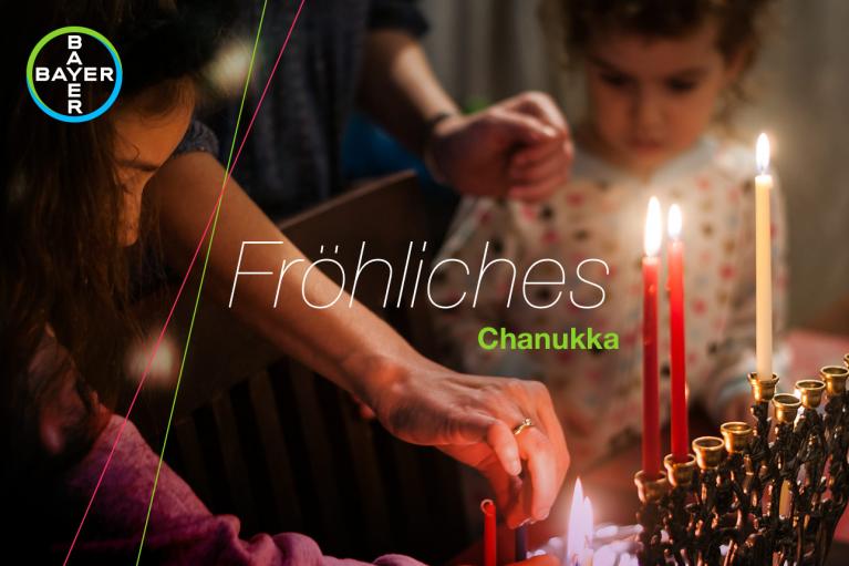 Two people light a menorah with several candles lit. Text reads "Fröhliches Chanukka" with a Bayer logo in the corner.