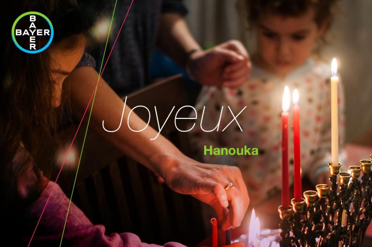 Adults and a child light a menorah for Hanukkah. The image features the words "Joyeux Hanouka" and the Bayer logo.