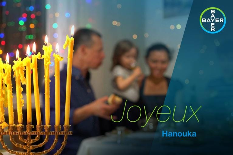 A family sits at a table with lit candles in the foreground. The image has text that reads "Joyeux Hanouka," alongside the Bayer logo.