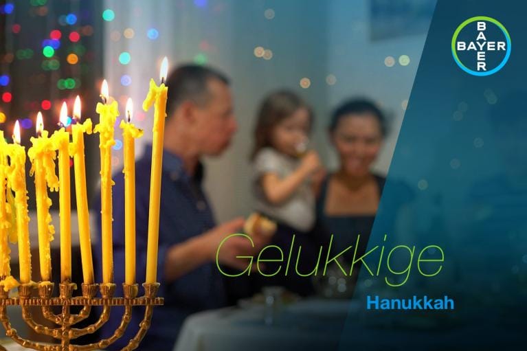 A lit menorah is in the foreground, with a family blurred in the background. Text reads "Gelukkige Hanukkah.