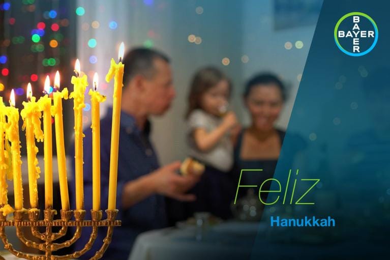 A menorah with lit candles is in the foreground; a family is gathered in the background celebrating Hanukkah. Text reads "Feliz Hanukkah.