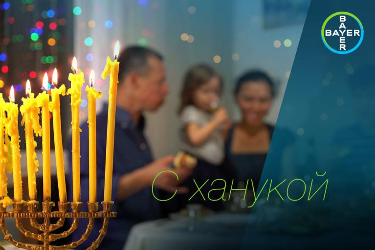 Lit menorah in the foreground with a blurred family having a meal in the background. Text in Russian reads "С ханукой" alongside Bayer logo.
