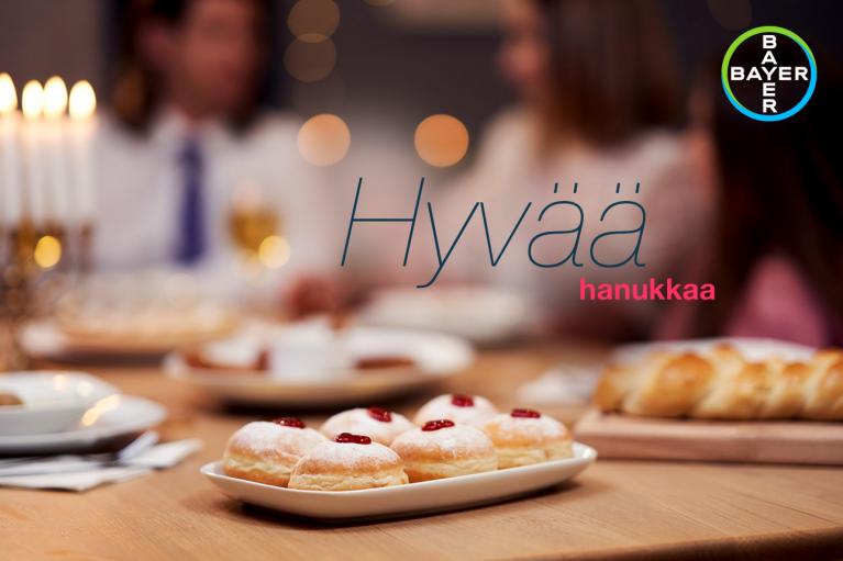 A table with pastries and bread, featuring text in Finnish "Hyvää hanukkah," with the Bayer logo in the corner.