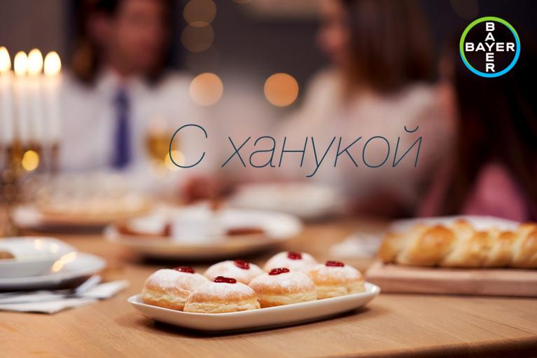 A plate of jelly-filled pastries on a table set for Hanukkah, with blurred people in the background and a Bayer logo in the corner.