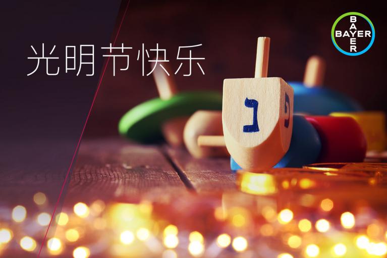 A dreidel with Hebrew letters surrounded by chocolate coins is on a wooden surface. The Bayer logo is in the top right corner, with Chinese text on the left.